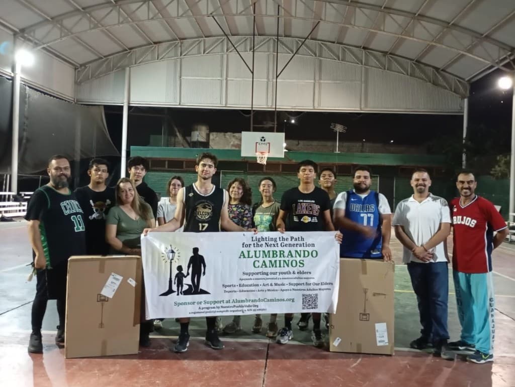 Youth, mentors, and families on a basketball court at night with the Alumbrando Caminos program banner — a Nuestro Pueblo Valle initiative.