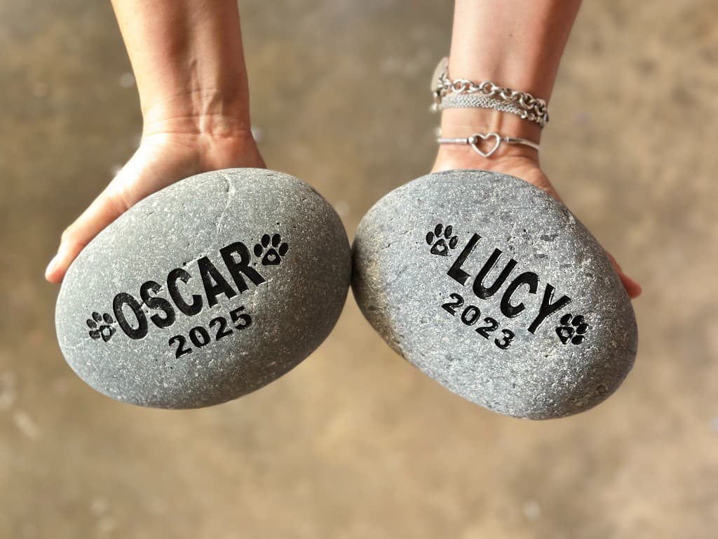 Hand-held engraved river stones honoring two pets — an example of the thoughtful keepsakes our brands help families create.